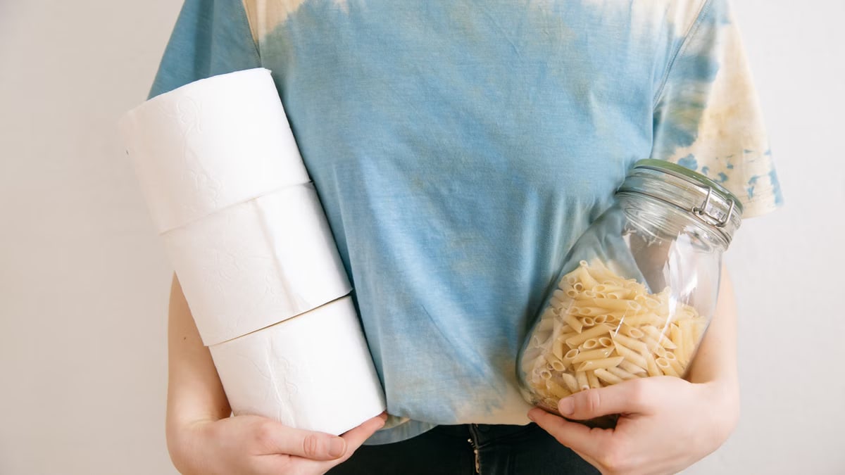 Woman in tie-dye shirt holding toilet paper rolls and pasta jar indoors.